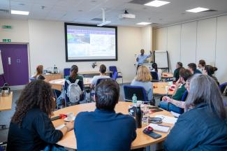 Lots of people sat on different round tables all facing someone at a lectern presenting with their arms out wide.  On the screen is a slide about Marek's disease
