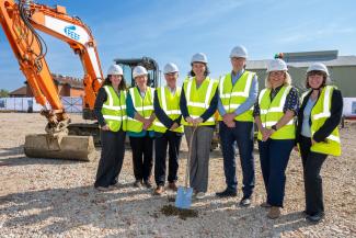 Seven people in a row wearing reflective jackets and hardhats. Laura Dance in the middle is holding a shovel. Behind the group are orange construction vehicles