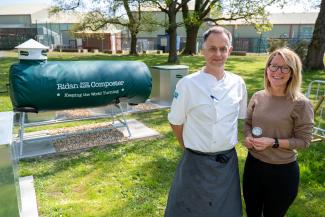 A Chef and a person stood on grass together looking at the camera. Behind them on the left is a green cylinder with metal legs. White words on the green composter read "ridan food waste composter. keeping the world turning."  