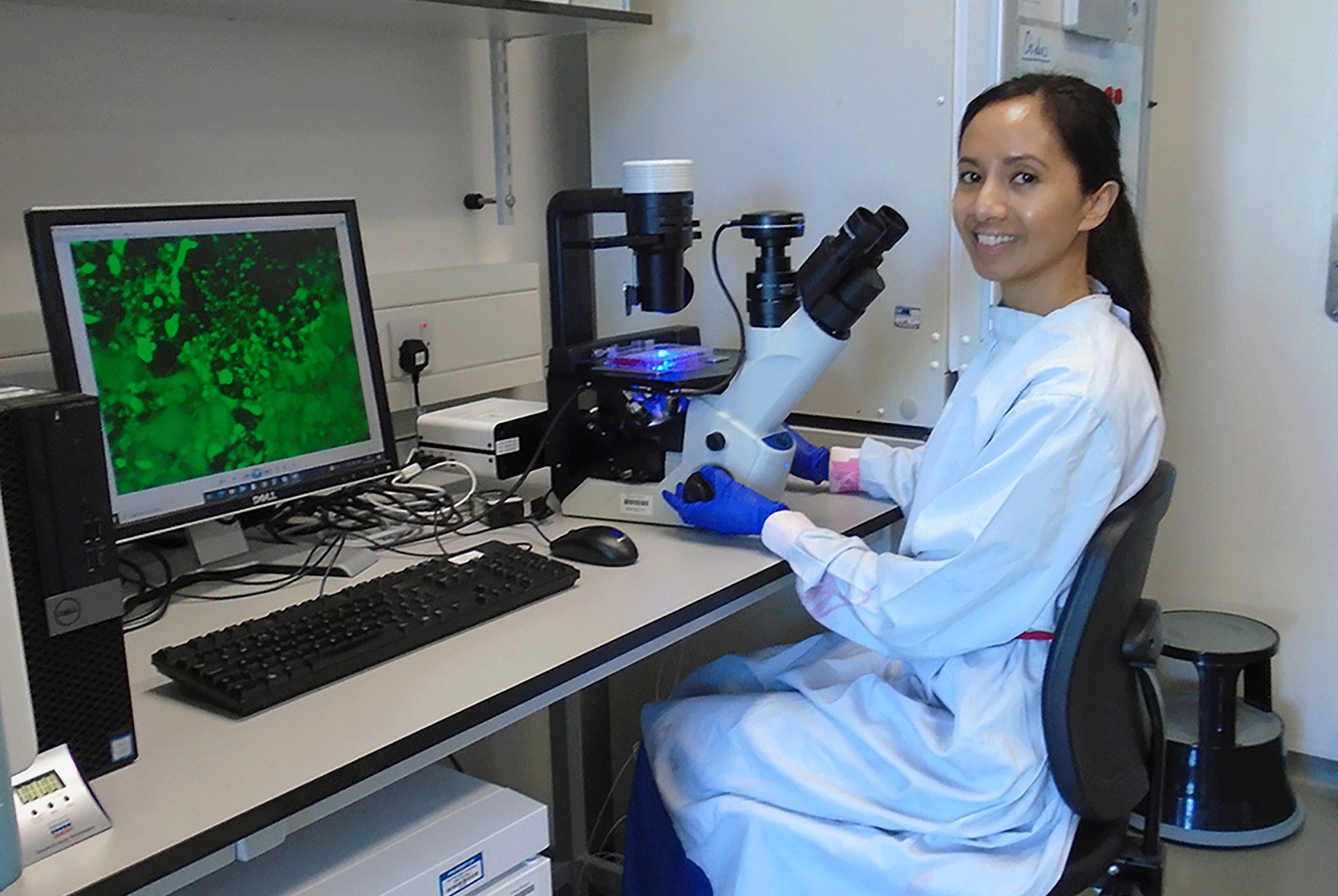 Amanda Corla in a lab coat and wearing purple gloves, sat on a chair in front of a microscope